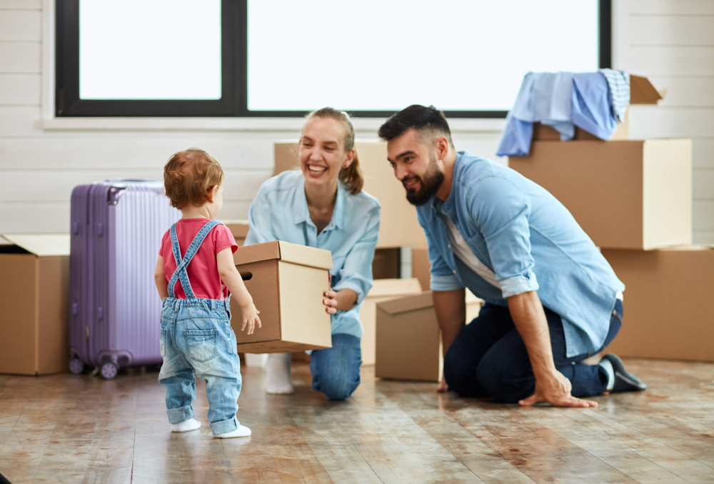 Dark-haired bearded man and fair-haired woman wear blue shirt sit on floor, smile and hold box that child brought. The background moving boxes and suit