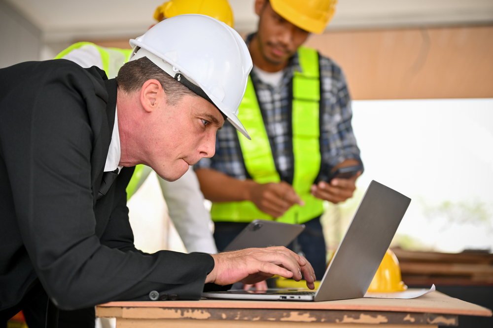 Side view of a determined and professional Caucasian businessman in a white hardhat and business suit using a laptop while working with a team of construction engineers at the construction site.