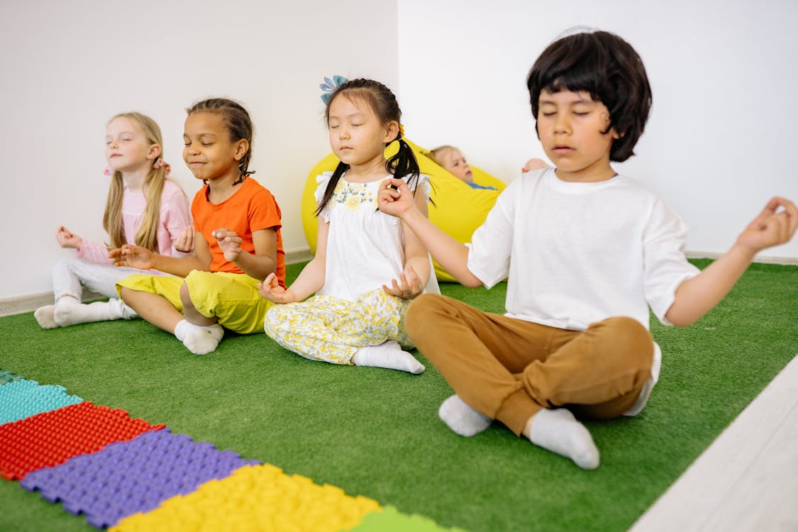 Free Group of diverse young children meditating in yoga poses on a green mat indoors. Stock Photo