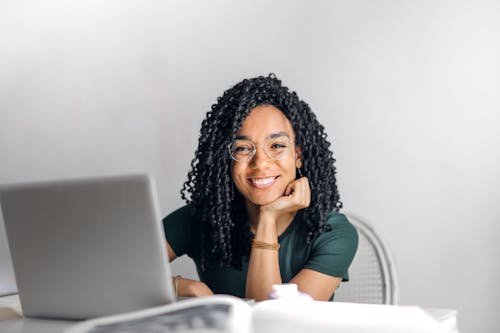 Free Joyful businesswoman with curly hair smiling at camera while using laptop indoors. Stock Photo