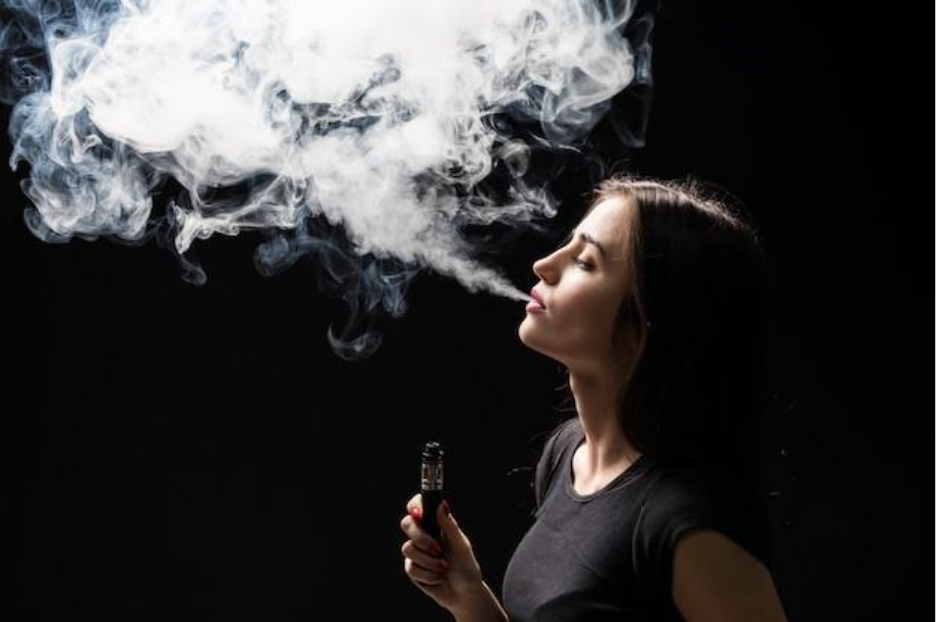young beautiful brunette woman smoking, vaping e-cigarette with smoke on the black wall