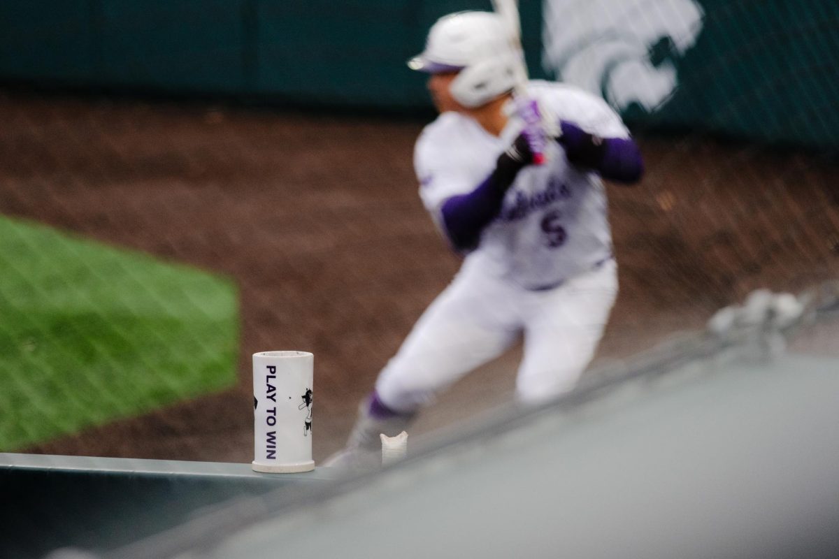 A bat weight states, "Play to Win" as a K-State Baseball player warms up in the background during the April 3, 2026 game against TCU at Tointon Family Stadium.