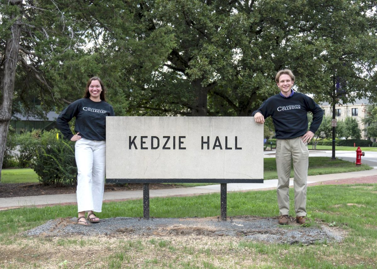 Kansas State Collegian Editor-in-Chief Greta Klein and Managing Editor Declan Phipps smile and pose with the Kedzie Hall sign in Kansas State Collegian sweatshirts. 