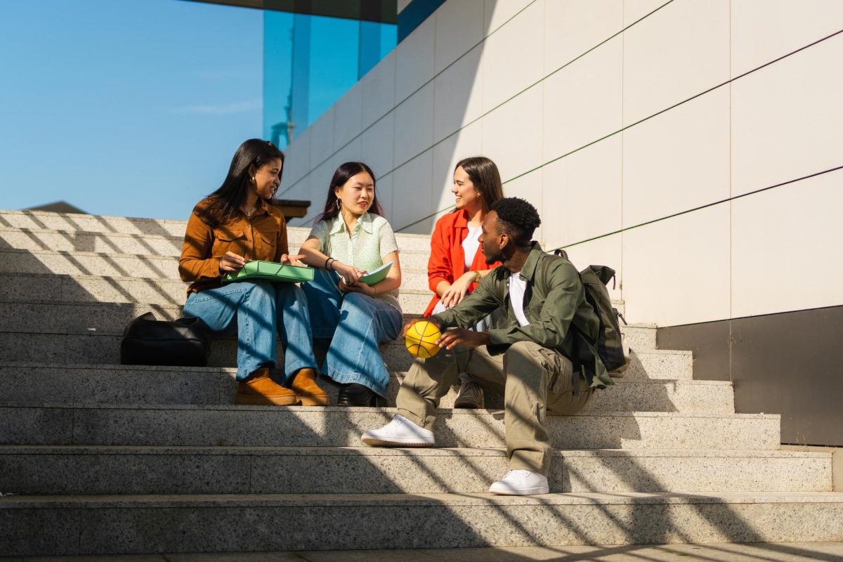 Diverse young adult friends sitting on concrete stairs, talking and smiling while holding books on a college campus