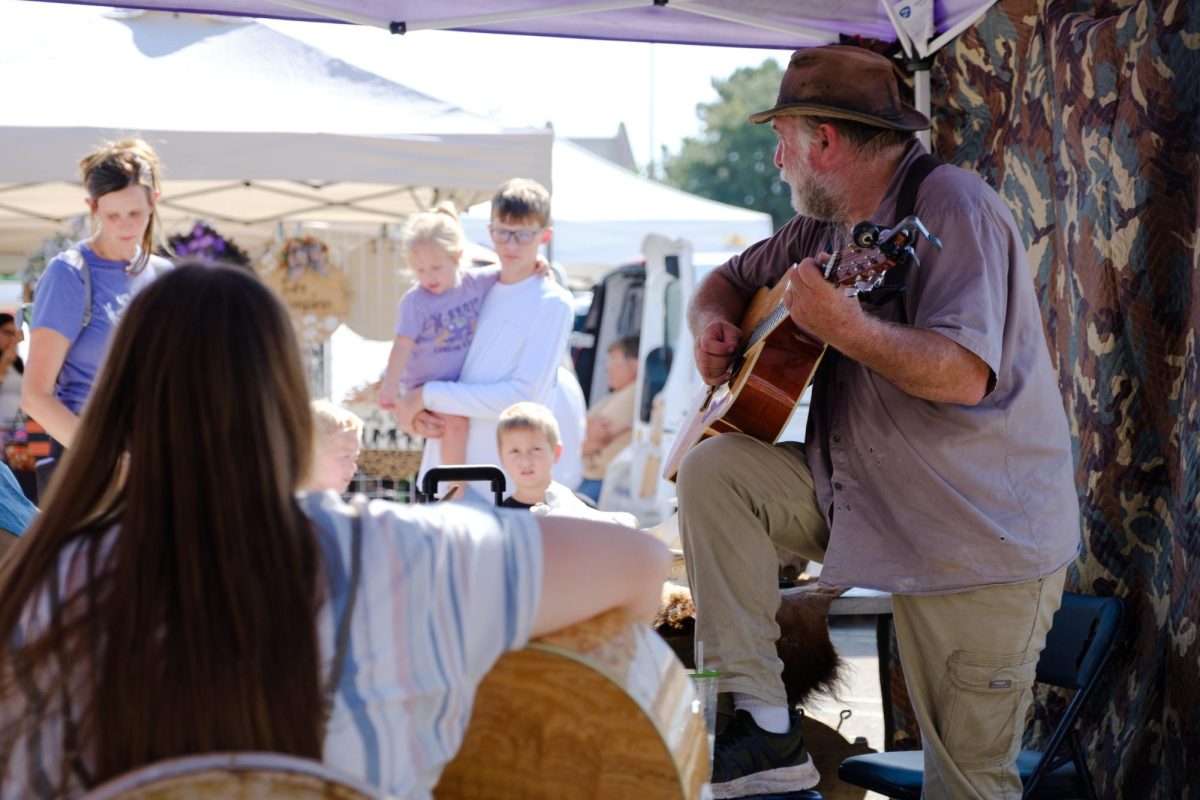 George LeRoux, a musician and vendor at the Manhattan Farmer's Market, plays guitar as he talks with a family of customers. LeRoux hosts open jam sessions alongside his vending booth, the Flint Hills Prairie Bison Preserve. 