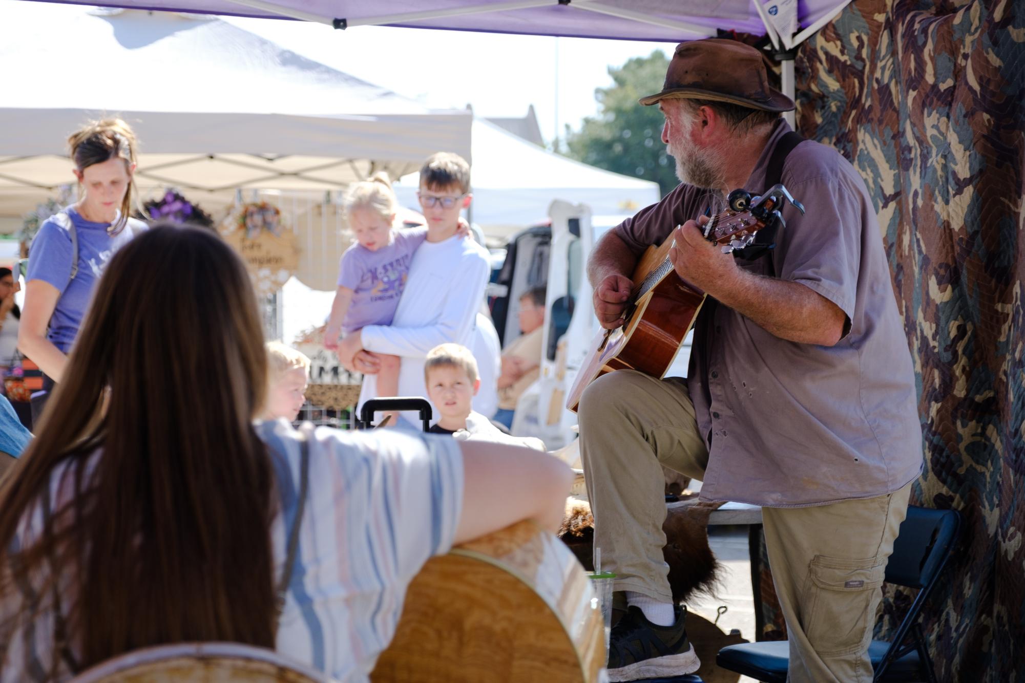 George LeRoux, a musician and vendor at the Manhattan Farmer's Market, plays guitar as he talks with a family of customers. LeRoux hosts open jam sessions alongside his vending booth, the Flint Hills Prairie Bison Preserve. 