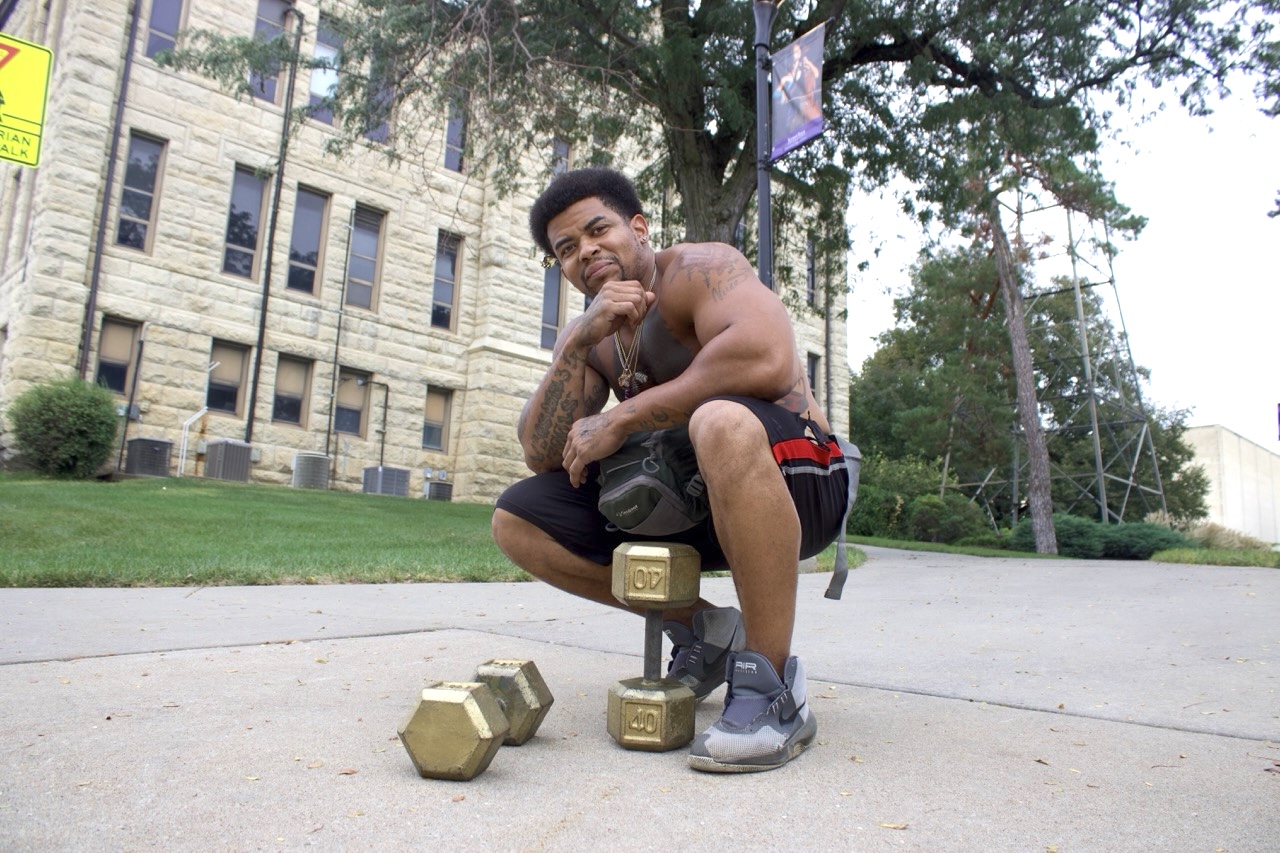Bryan Vereen crouches and poses with his iconic dumbbells on K-State's campus. Vereen is known for walking around Manhattan and curling gold dumbbells down Anderson Avenue.